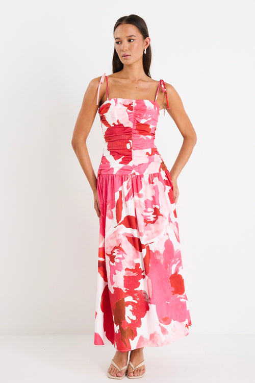 Woman wearing a floral pink and red ruched midi dress with tie-up straps and a flared hem, styled with minimal sandals.