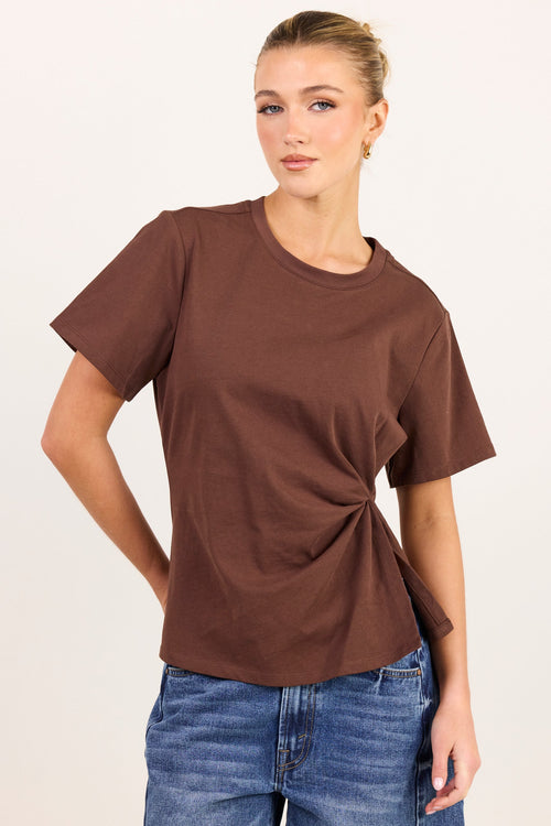 A woman modeling a brown short-sleeve t-shirt with a twisted side detail, paired with blue jeans, against a plain off-white background.
