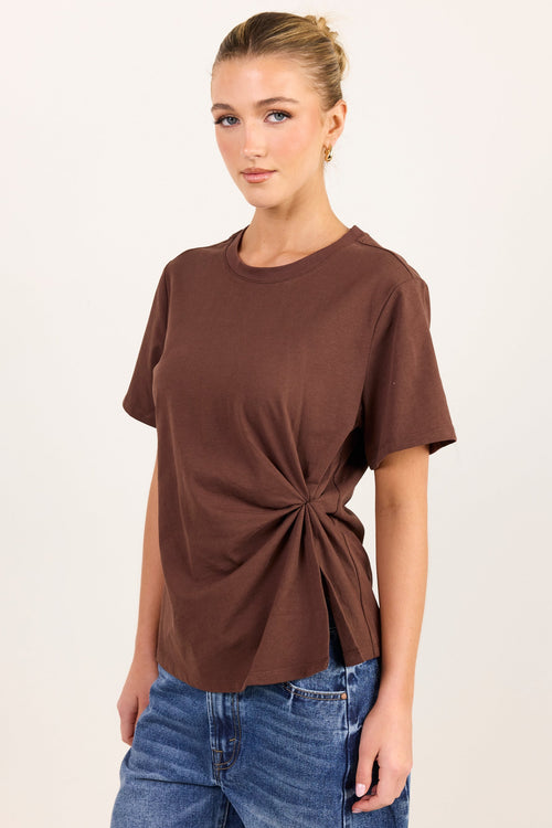 A woman modeling a brown short-sleeve t-shirt with a twisted knot detail at the side, paired with blue jeans, against a plain off-white background.