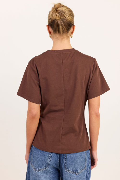 A woman viewed from behind wearing a brown short-sleeve t-shirt and blue jeans, standing against a plain off-white background.