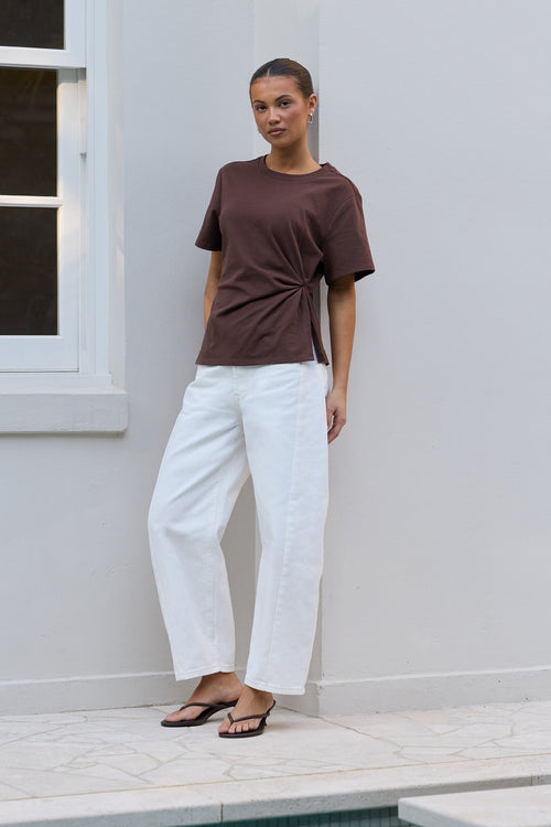 A woman stands outdoors against a white wall next to a pool, wearing a brown short-sleeve t-shirt with a twisted side detail and white wide-leg pants.