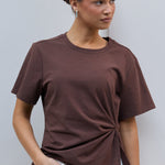 A woman wearing a brown short-sleeve t-shirt with a twisted knot detail at the hem, paired with white pants, standing against a light gray wall.