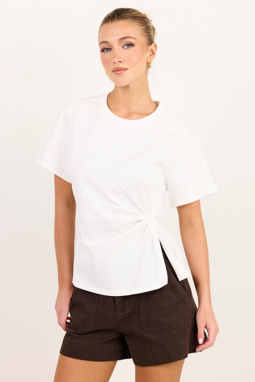 A woman modeling a white short-sleeve t-shirt with a twisted knot detail at the side hem, paired with dark brown shorts, against a plain off-white studio background.