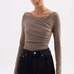 A woman modeling a taupe-brown long-sleeve top with an asymmetrical draped neckline, paired with black jeans, against a plain light gray background.