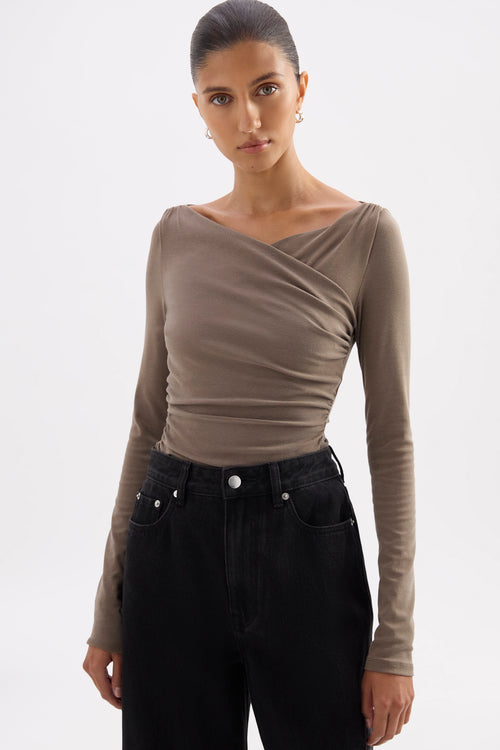 A woman modeling a taupe-brown long-sleeve top with an asymmetrical draped neckline, paired with black jeans, against a plain light gray background.