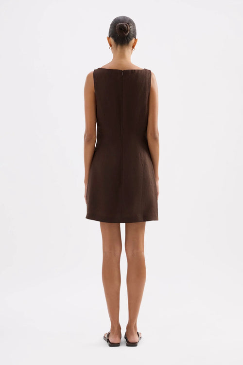 A woman viewed from the back wearing a sleeveless brown mini dress with a straight neckline and a simple dark heel, standing against a plain white studio background.