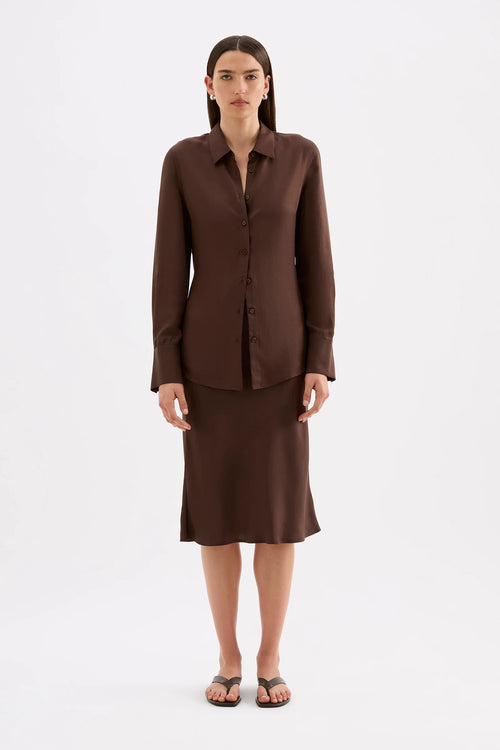 A woman stands against a plain light gray studio background wearing a matching dark brown button-front blouse and knee-length skirt set with black flip-flop sandals.