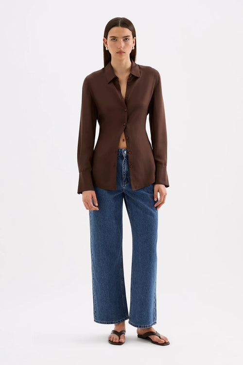 A model stands against a light gray studio background wearing a dark brown long-sleeve button-front shirt, blue wide-leg jeans, and brown sandals.