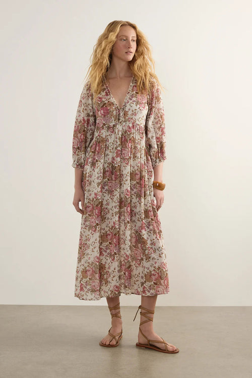 A woman modeling a long-sleeved midi dress with a pink and green floral pattern on a cream background, paired with brown strappy flat sandals, set against a neutral studio backdrop.