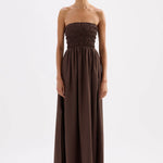 A woman modeling a strapless, smocked-bust maxi dress in a solid dark brown color, standing against a plain white studio background.