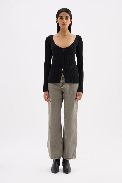 A model stands against a plain white studio background wearing a black long-sleeve knit top with a wide scoop neckline and front buttons, paired with light gray wide-leg pants and black ankle boots.