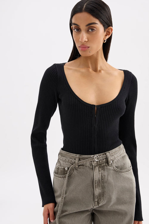 A woman wearing a black ribbed long-sleeve scoop-neck top with a front zipper, paired with gray high-waisted jeans, against a plain light gray background.