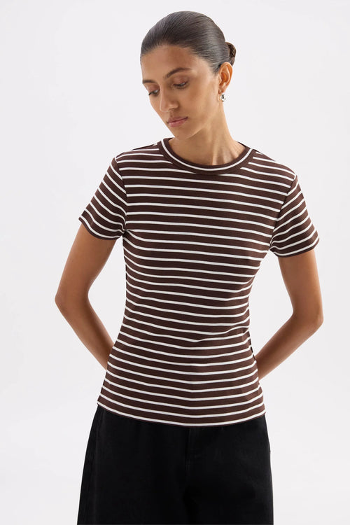 A woman wearing a fitted short-sleeve crewneck top with thin horizontal brown and white stripes, paired with black pants, photographed against a light gray studio background.