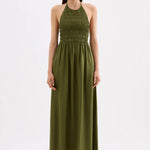 A woman modeling a sleeveless olive green halter maxi dress with a smocked bodice, standing against a plain white studio background.