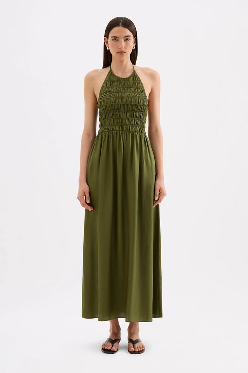 A woman modeling a sleeveless olive green halter maxi dress with a smocked bodice, standing against a plain white studio background.