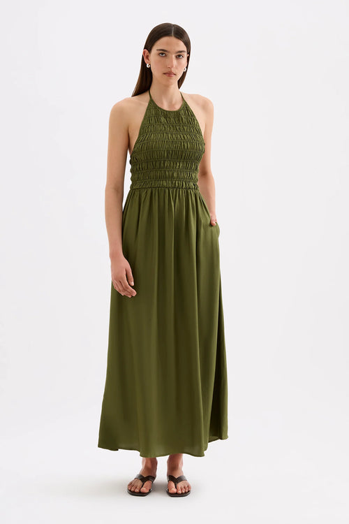 A woman modeling a sleeveless olive green halter maxi dress with a smocked bodice and side pockets, standing against a plain light gray studio background.