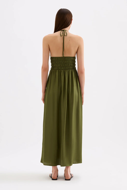 A woman viewed from the back wearing an olive green halter maxi dress with a smocked bodice and tie-back straps, standing against a plain white studio background.