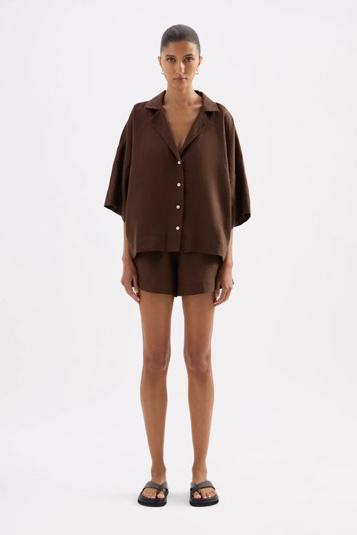 A model stands against a light gray studio background wearing a matching dark brown short-sleeve button-front shirt and shorts set, with black slide sandals.