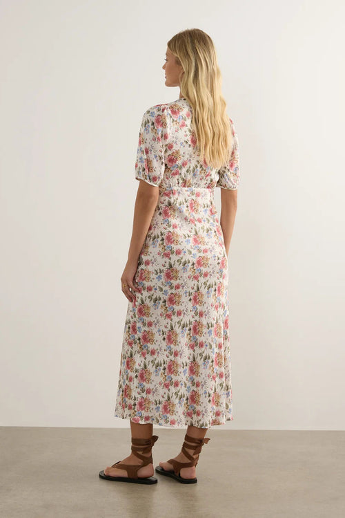 A woman stands with her back to the camera wearing a short-sleeve midi dress with a pink, red, and green floral pattern on a cream background. She is wearing brown strappy sandals in a minimalist studio setting with a light beige wall and concrete floor.