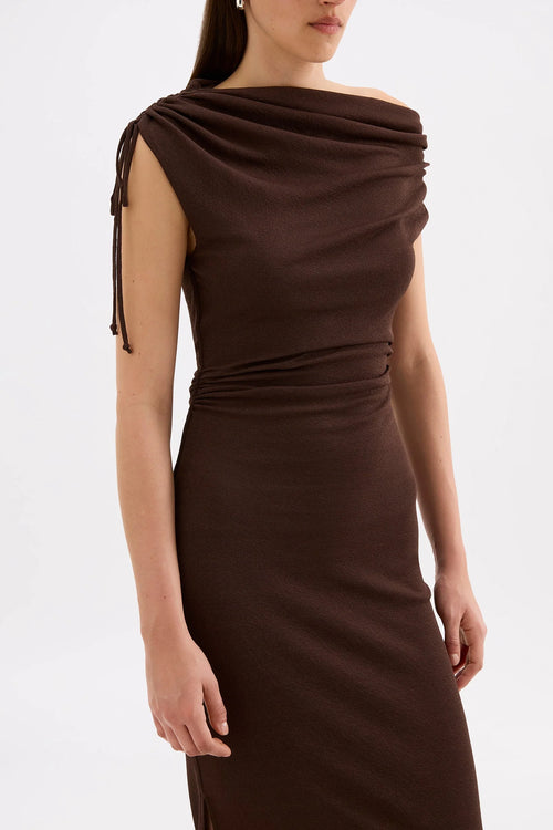 A woman wearing a dark brown off-the-shoulder knit dress with side tie details and a ruched waist, shown against a plain light gray studio background.