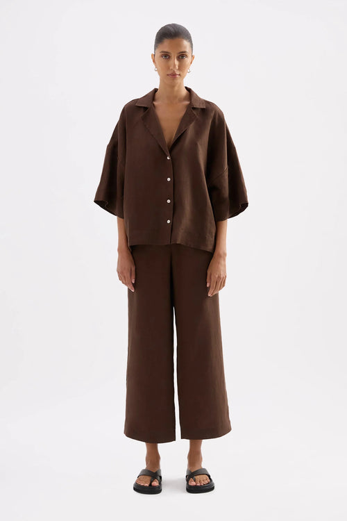 A model stands against a light gray studio background wearing a matching dark brown short-sleeve button-front shirt and wide-leg cropped pants set, paired with black sandals.