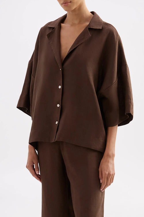 A model wearing a dark brown short-sleeve button-up shirt and matching brown trousers against a plain white studio background.