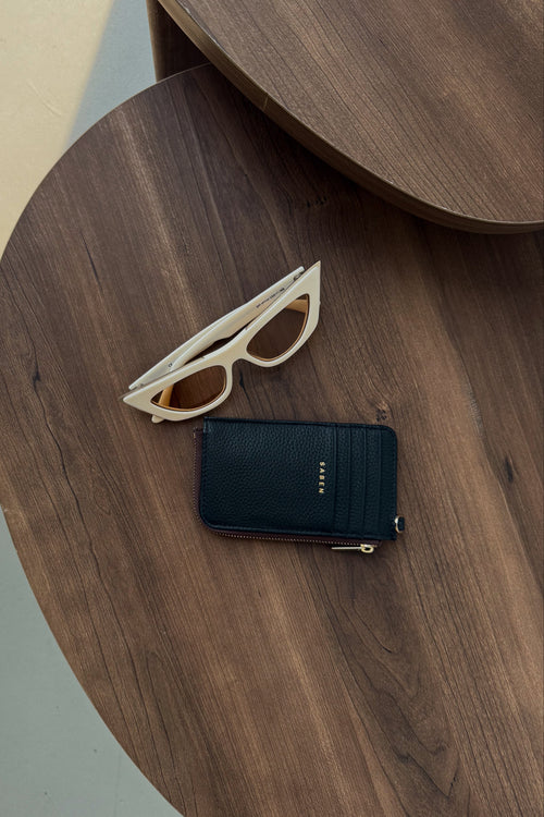 A navy blue leather zip wallet with gold hardware lies on a dark wood table next to a pair of cream-colored sunglasses with amber lenses.