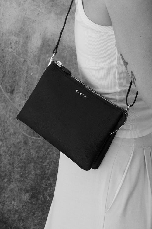 A black leather crossbody bag with a silver-tone zipper and a small 'FREDERIC T' logo on the front, worn by a model in a white dress against a textured concrete wall.