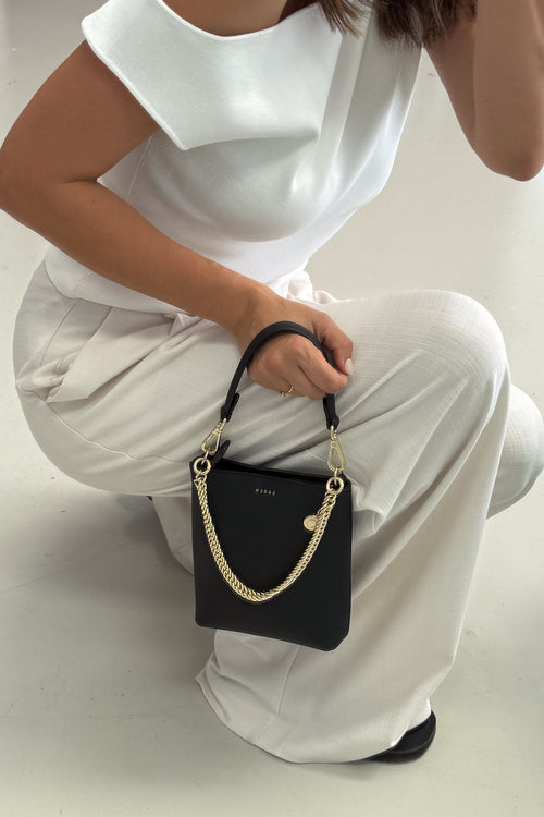 A woman in a white outfit holds a black GUESS bucket bag with a gold chain strap and gold logo detail on a light gray floor.