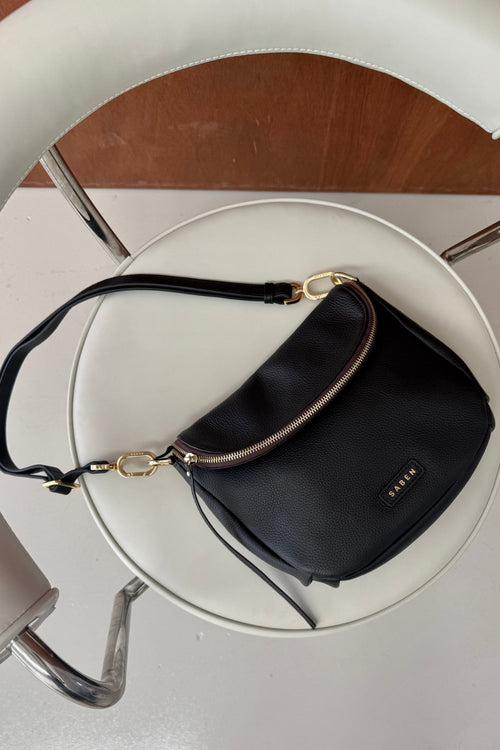 A black leather shoulder bag with gold-tone hardware and a small 'FASHION' logo plaque, resting on a white circular chair.