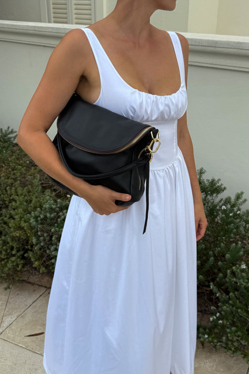 A woman wearing a white sleeveless midi dress and holding a black leather shoulder bag with gold hardware, standing outdoors in front of a light-colored wall and greenery.