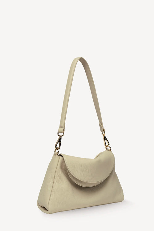 A light beige leather shoulder bag with a curved flap and a long shoulder strap, shown from a three-quarters angle on a white background.