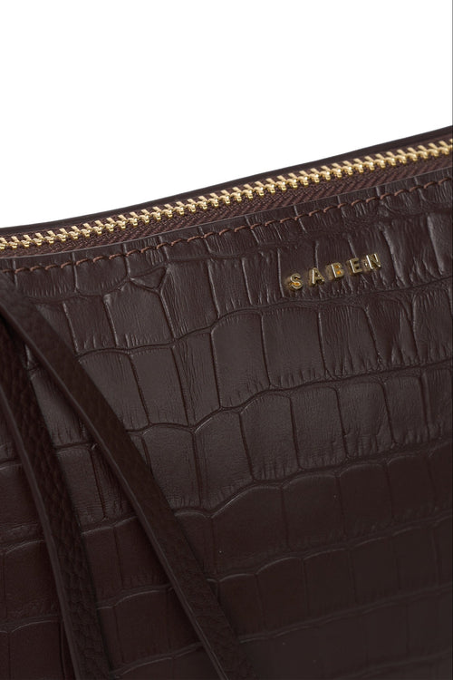 Close-up of a dark brown crocodile-embossed leather bag with a gold zipper and gold SABEN logo on a white background.
