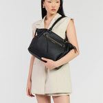 A model wearing a beige sleeveless blazer dress holds a black leather shoulder bag with gold hardware against a light gray studio background.