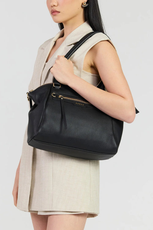 A woman wearing a beige sleeveless blazer and matching skirt carries a black leather shoulder bag with gold hardware and a visible 'GUESS' logo on the front, against a light gray studio background.