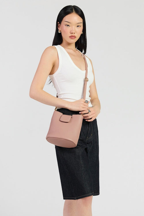 A woman modeling a dusty pink leather shoulder bag with a matching strap, styled with a white tank top, black skirt, and black sandals against a light gray studio background.