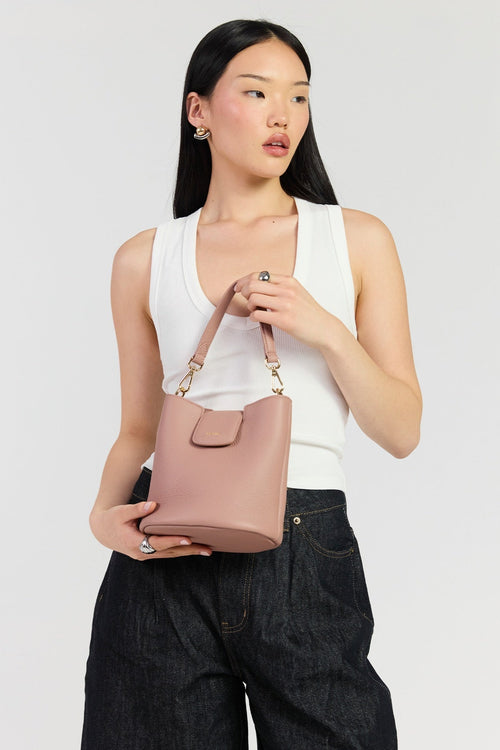 A model wearing a white tank top and dark wide-leg jeans holds a dusty pink leather bucket bag with gold-tone hardware against a light gray studio background.