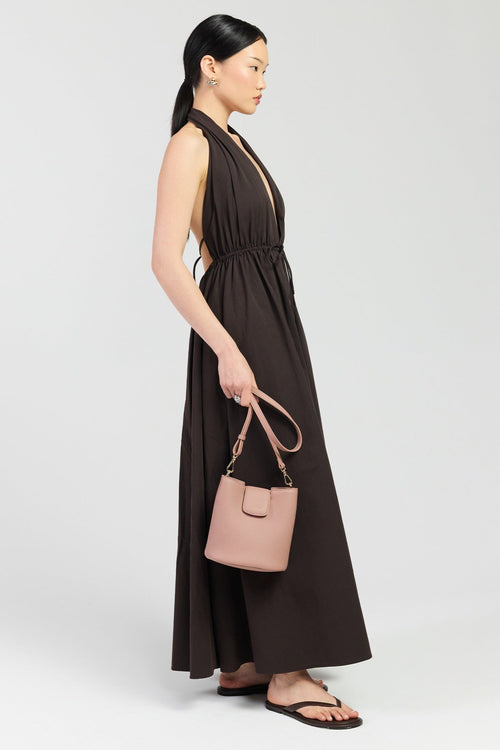 A woman stands in profile wearing a sleeveless dark brown maxi dress with a deep V-neckline and gathered waist, holding a dusty pink handbag. She is wearing dark brown flat sandals against a light gray studio background.