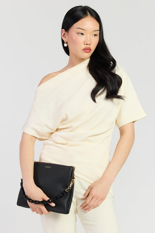A model wearing a cream-colored asymmetrical short-sleeve top and matching trousers, holding a black leather clutch bag with a gold chain strap against a light gray background.