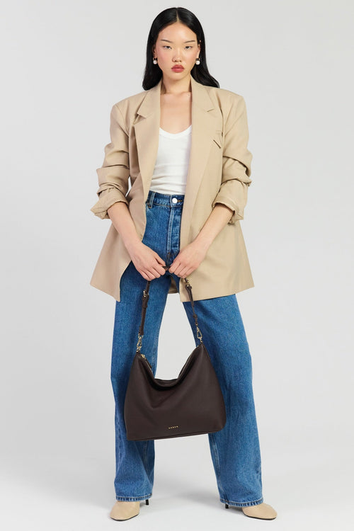 A model stands against a light gray studio background wearing a beige blazer over a white tank top, blue wide-leg jeans, and beige shoes, while holding a dark brown hobo shoulder bag.