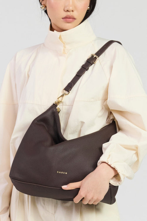 Person holding a dark brown shoulder bag with a visible brand logo against a neutral background