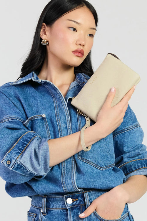 A woman wearing a blue denim shirt holds a beige leather clutch with a matching wrist strap against a light gray background.