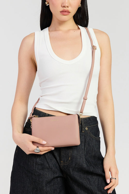 A model wearing a white ribbed tank top and dark denim jeans holds a dusty pink faux-leather crossbody bag with a matching shoulder strap against a light gray studio background.