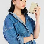 A model wearing an oversized blue denim jumpsuit holds a small beige leather clutch in front of her face, photographed against a solid light gray background.