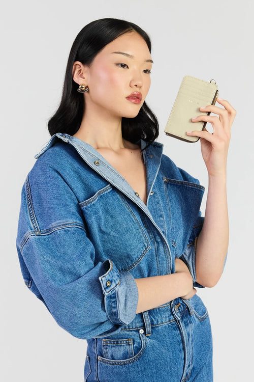 A model wearing an oversized blue denim jumpsuit holds a small beige leather clutch in front of her face, photographed against a solid light gray background.