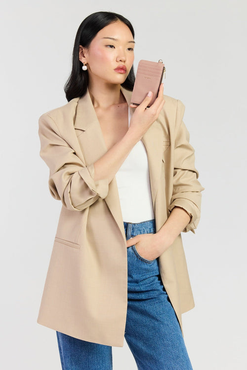 A woman modeling a beige oversized blazer over a white top and blue jeans, holding a pink phone case against a light gray studio background.