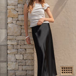 Woman standing against a stone wall wearing sunglasses and a white top with a black skirt.