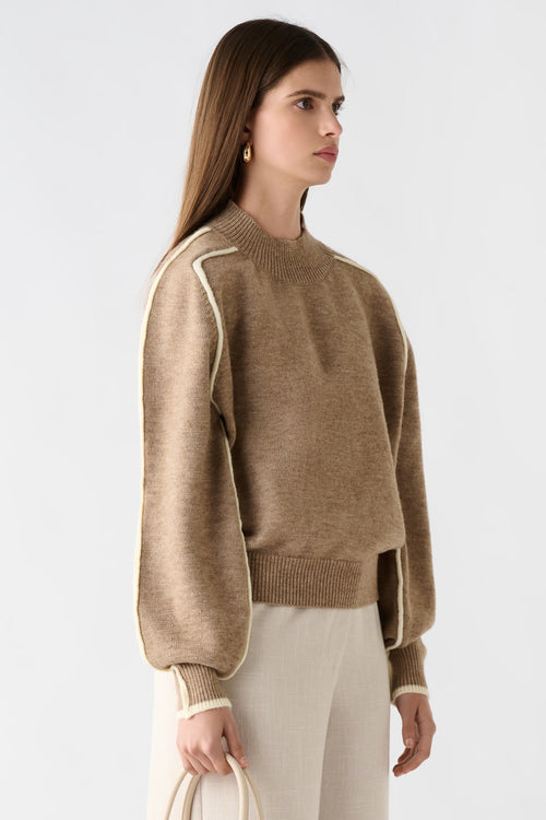 Woman wearing a beige sweater and white pants holding a cream handbag on a light gray background
