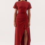 A woman modeling a rust-red midi dress with short flutter sleeves, a boat neckline, a ruched waist, and a high-low front slit, photographed against a seamless white studio background.