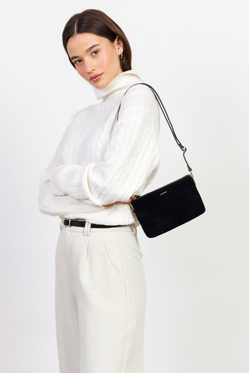 Woman in a white outfit holding a black handbag against a light gray background